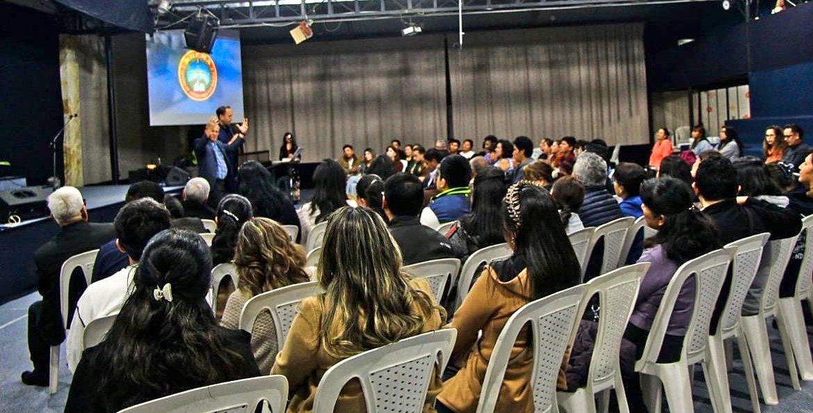 A full room of students and leaders listening as David teaches, the RHEMA Bolivia seal on the screen