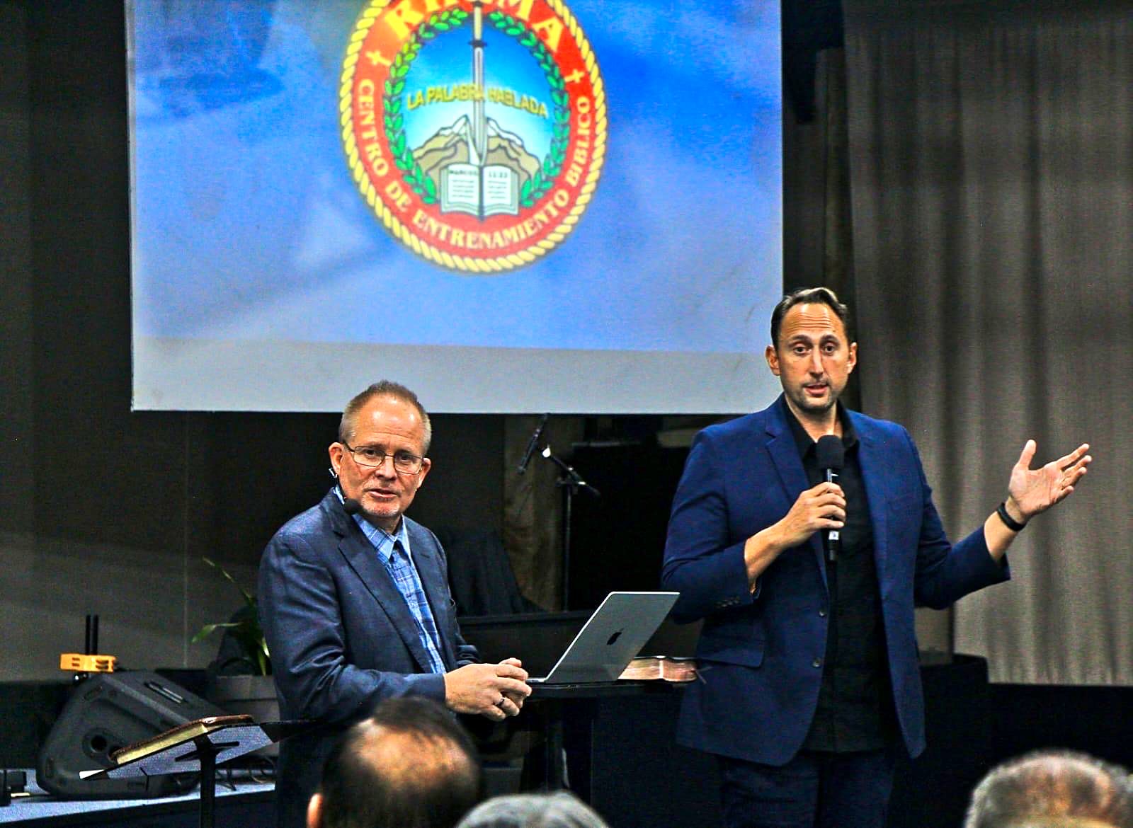 David Larson on stage with a guest minister, the RHEMA Bolivia seal on the screen behind them