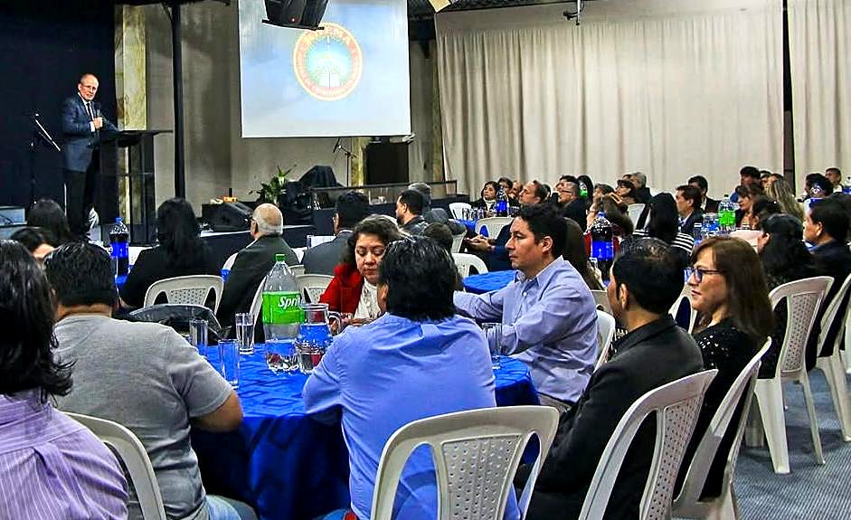 David Larson speaking from the podium to pastors and leaders seated at dinner tables, the RHEMA Bolivia seal on the screen